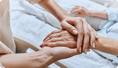 Close-up of a caregiver gently holding the hand of an elderly person lying in bed.