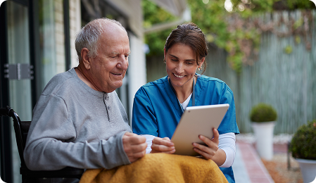 A healthcare worker assists an elderly man in a wheelchair, both engaged with a tablet in a comfortable outdoor setting.