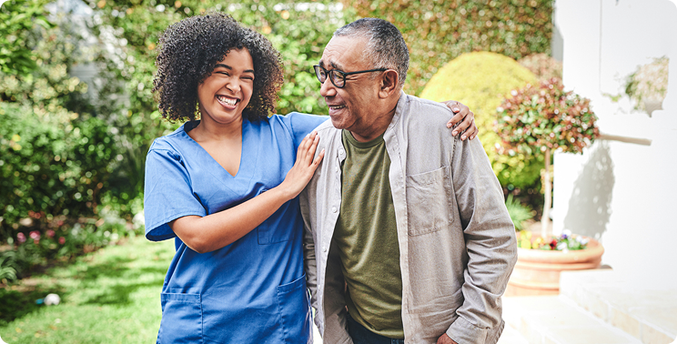 A smiling healthcare provider in scrubs interacts with a cheerful older man in a garden setting.