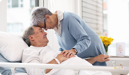 A woman affectionately leans her forehead against a man's in a hospital room, showcasing a moment of tenderness.