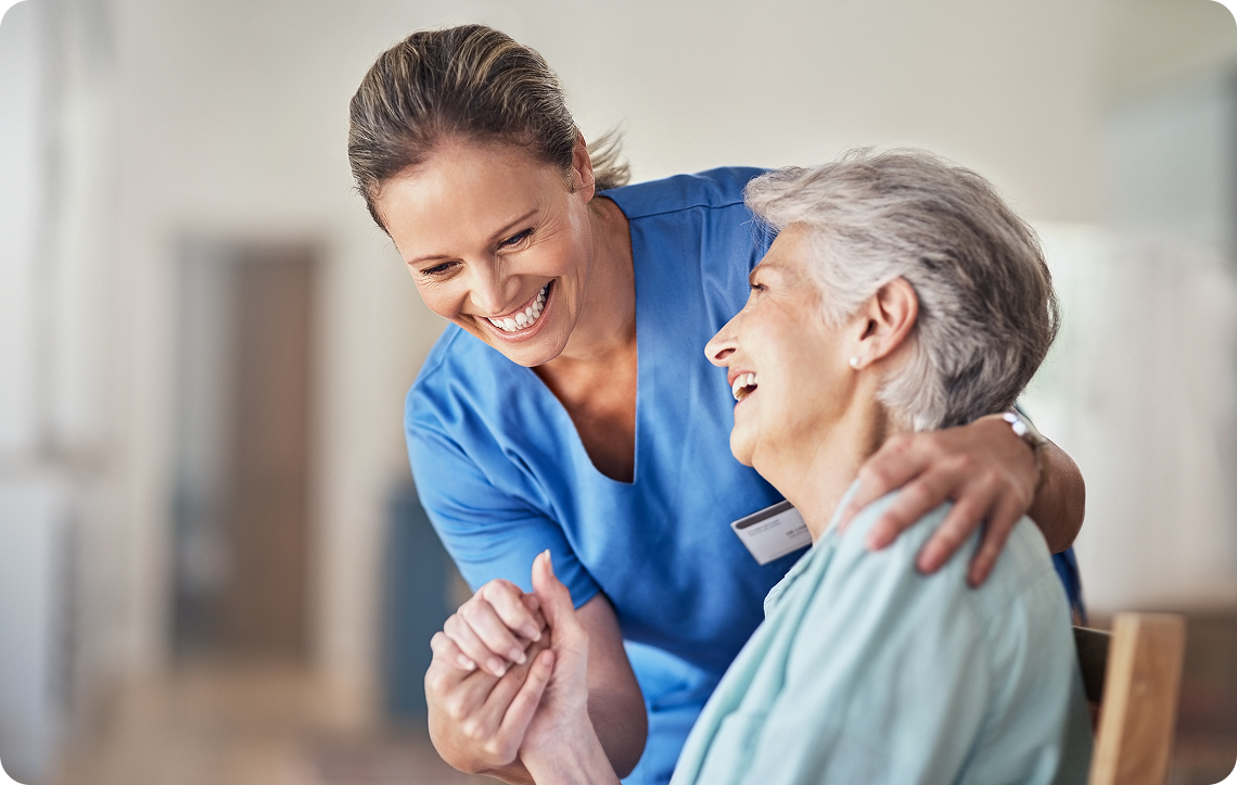 A female caregiver smiles and holds the hands of a laughing senior woman.