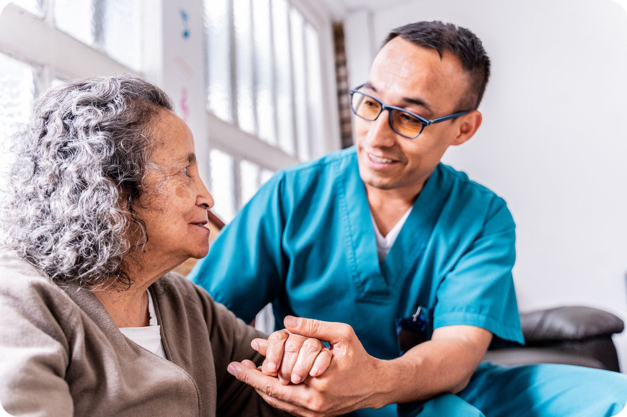 A kind male caregiver holds a senior woman's hands as they smile and talk together.
