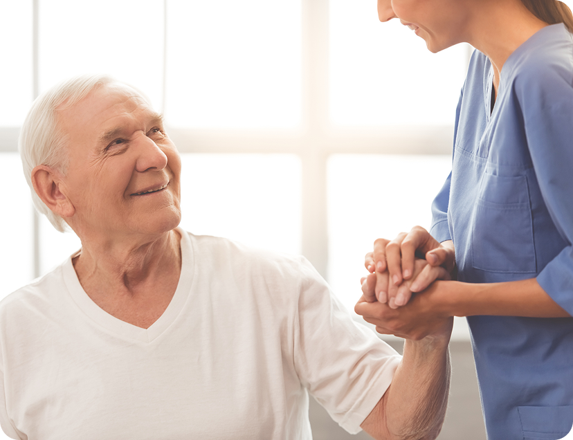 An elderly man smiling warmly as he holds hands with a female healthcare worker in blue scrubs, who is leaning slightly toward him in a caring gesture.