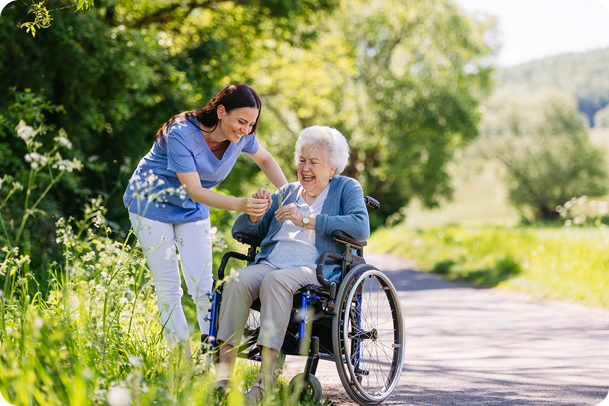 A female caregiver in scrubs smiles at a happy senior woman in a wheelchair on a path
