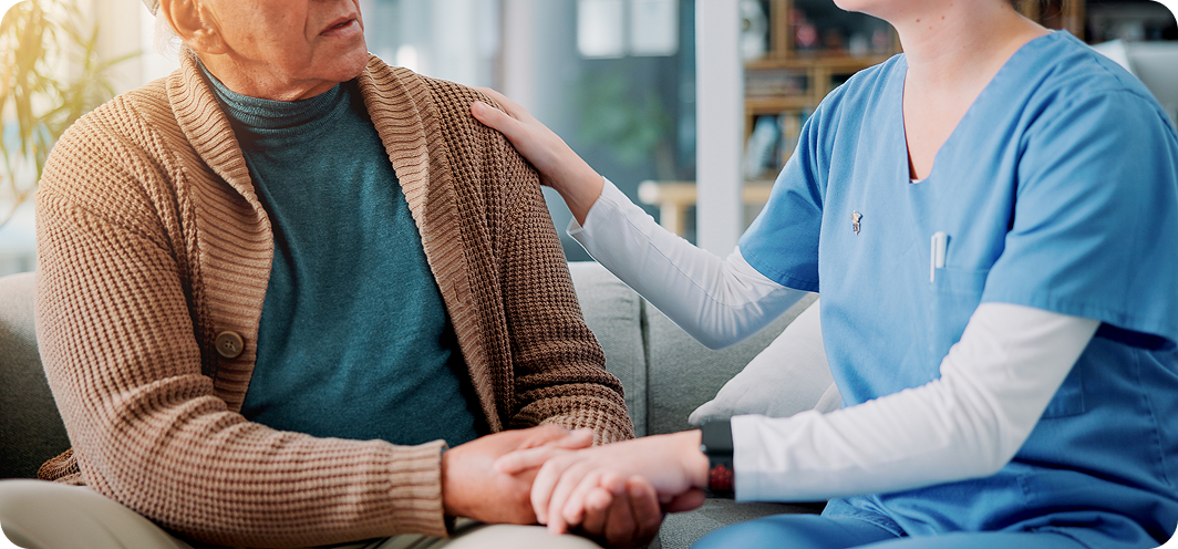 A caring female caregiver holds a senior man's hand and rests her other hand on his shoulder.