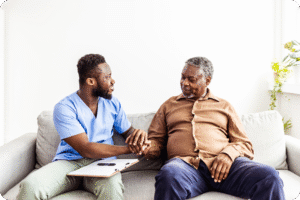 A friendly male caregiver holds the hand of a senior man while they sit on a couch together.