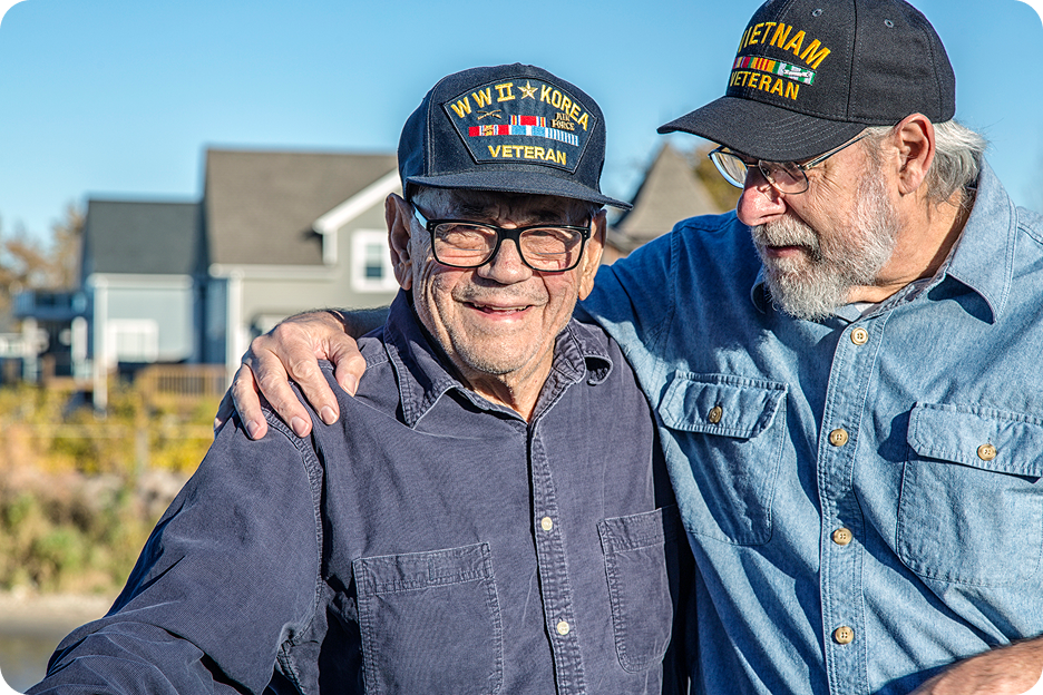 Two male senior veterans wearing a cup and standing with their arms around each other outside on a sunny day.