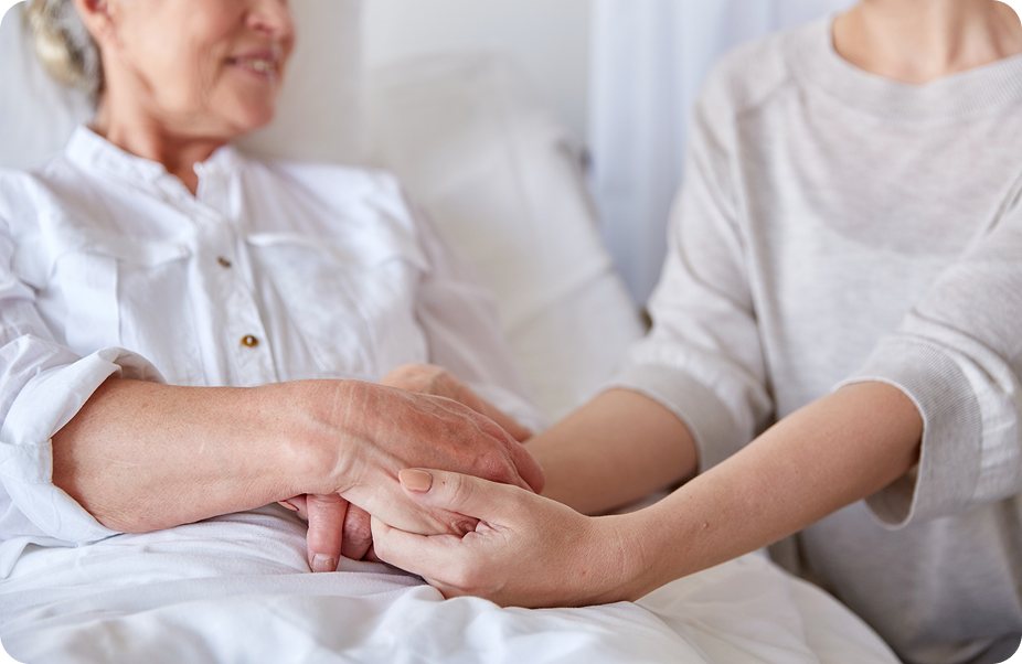 A caregiver gently holds the hands of a senior woman in a hospital bed.