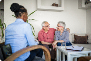A female caregiver sits across from a happy senior couple, with her arm around her partner.