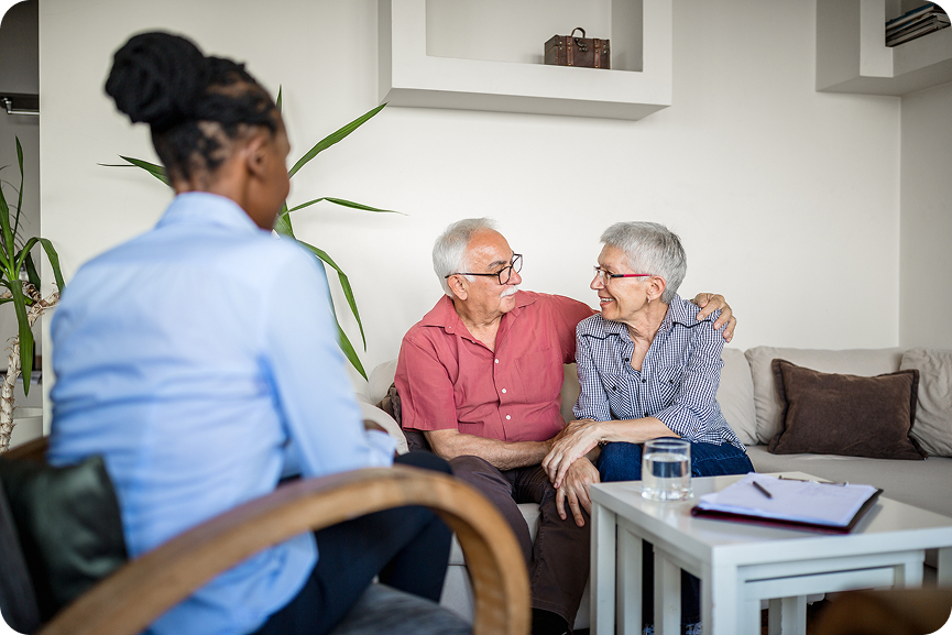 A female caregiver sits across from a happy senior couple, with her arm around her partner.