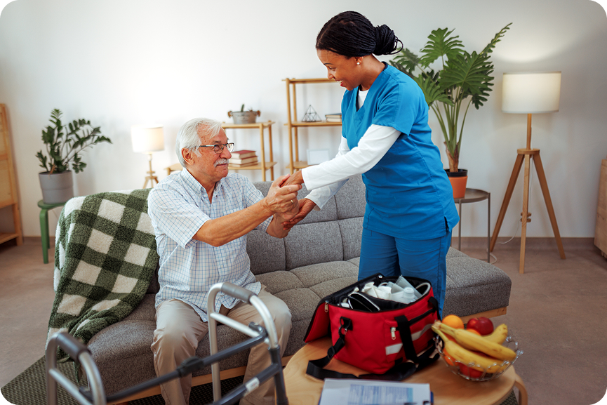 A friendly female caregiver helps a senior man with his medication in a living room, with a walker and a medical bag nearby.