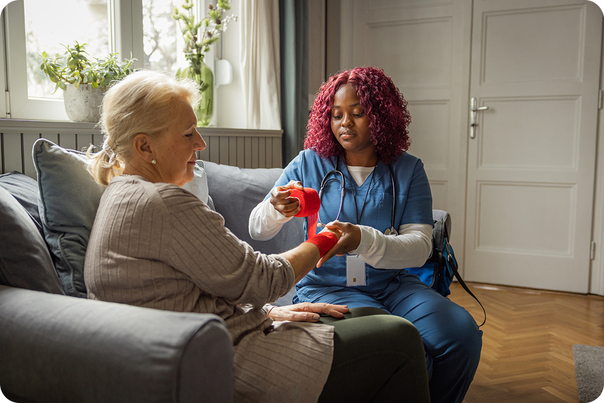 A smiling caregiver helps a senior woman with red hand weights during physical therapy at home.