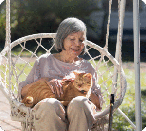 A senior woman sits on a swing chair and pets a cat on her lap.