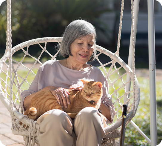 A senior woman sits on a swing chair and pets a cat on her lap.