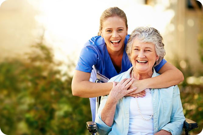 A cheerful female caregiver smiles with her arms around a happy senior woman in a wheelchair outdoors.