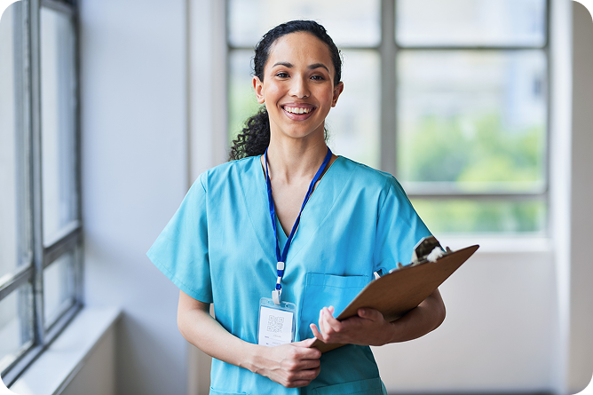 A female nurse in blue scrubs smiles at the camera while holding a clipboard in a hallway.