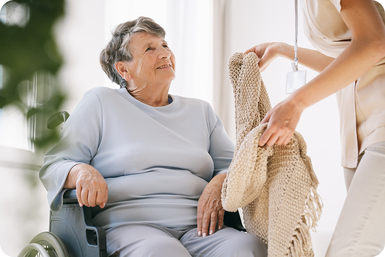 A caregiver helps a senior woman in a wheelchair by giving her a blanket.