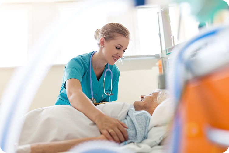A female nurse with a stethoscope smiles at a senior patient in a hospital bed.
