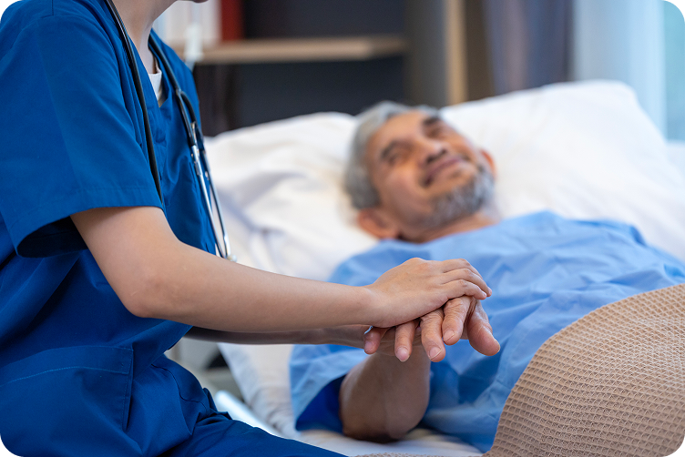 A caregiver holds the hand of a senior man lying in a hospital bed.