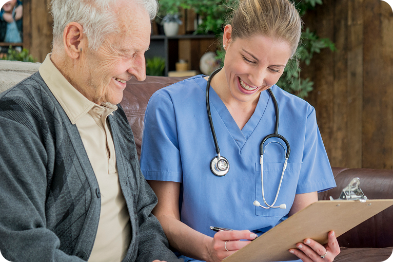 A smiling female caregiver holds a clipboard and talks with a senior man on a couch.