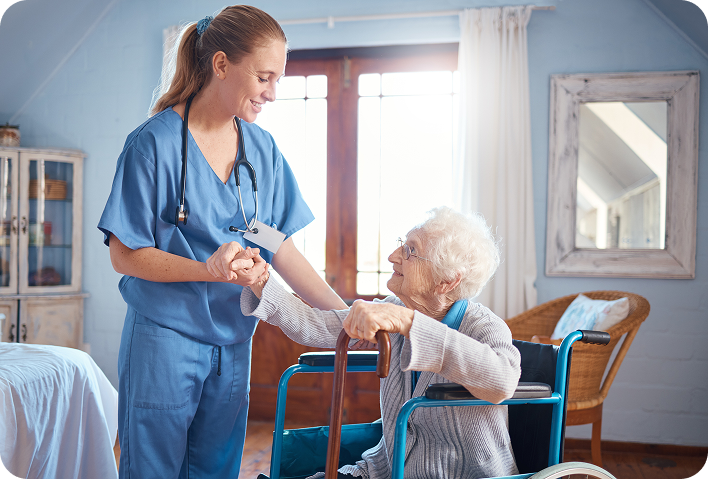 A smiling young female caregiver in blue scrubs stands next to a happy elderly woman in a wheelchair, holding her hand and providing comfort and assistance in a bright room.