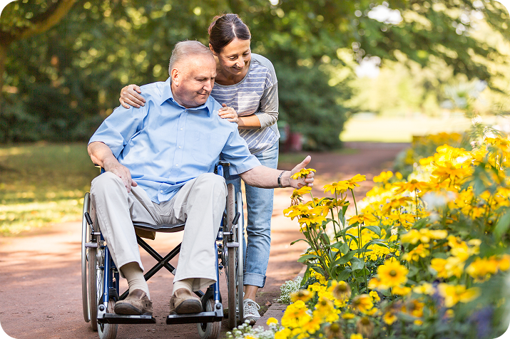 A man in a wheelchair and his caregiver enjoy a park, with the man reaching for a yellow flower.