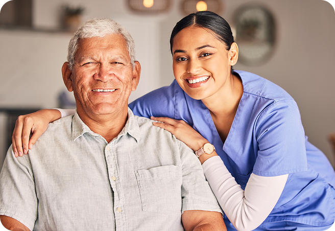 A young female caregiver in blue scrubs smiles with her arm around a happy senior man.