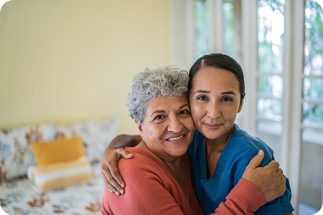 A smiling young caregiver in blue scrubs and a joyful senior woman with gray hair embrace, looking directly at the camera with a warm, happy expression.