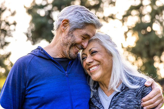A happy senior couple embraces outdoors. The man, with his arm around the woman, smiles while touching foreheads, as the woman leans into him and smiles happily.