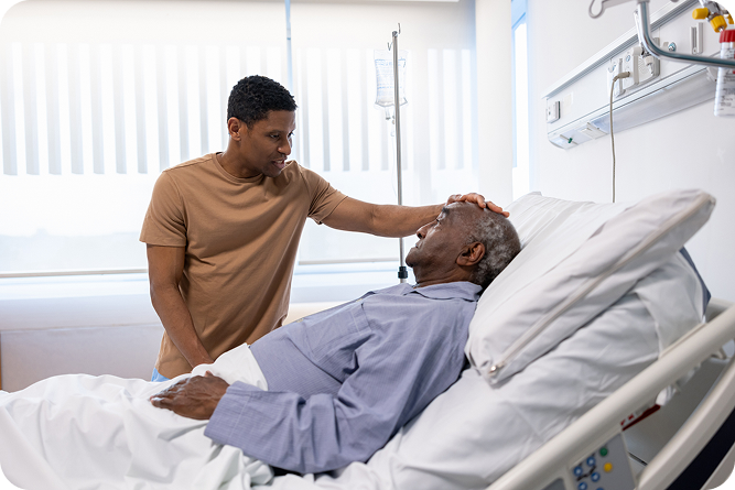A younger man stands beside a hospital bed and gently touches the forehead of an older man lying down, showing care and concern.