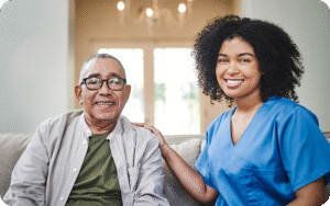 A happy young female caregiver smiles at the camera with her hand on the shoulder of a cheerful elderly man, as they sit together on a couch.