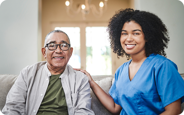 A happy young female caregiver smiles at the camera with her hand on the shoulder of a cheerful elderly man, as they sit together on a couch.