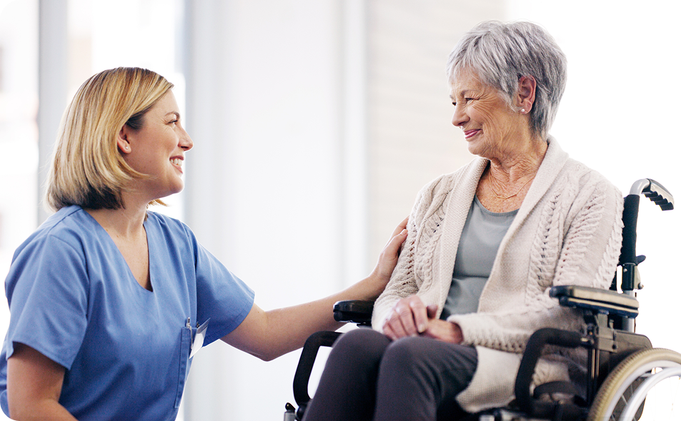 A caring female caregiver in blue scrubs smiles at a happy senior woman in a wheelchair, with her hand gently on the woman's shoulder as they share a warm moment.
