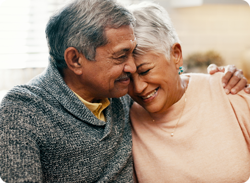 A loving senior couple smiles and presses their heads together, with the man's arm around the woman's shoulder, conveying a sense of warmth and affection.