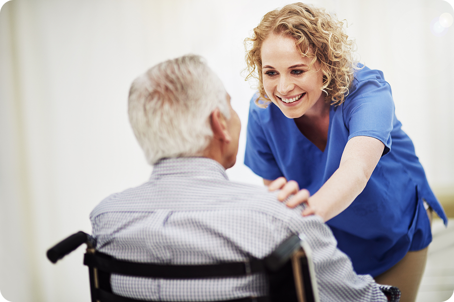 A friendly female caregiver in blue scrubs smiles at a senior man in a wheelchair, with her hand gently on his shoulder, as they share a warm conversation.