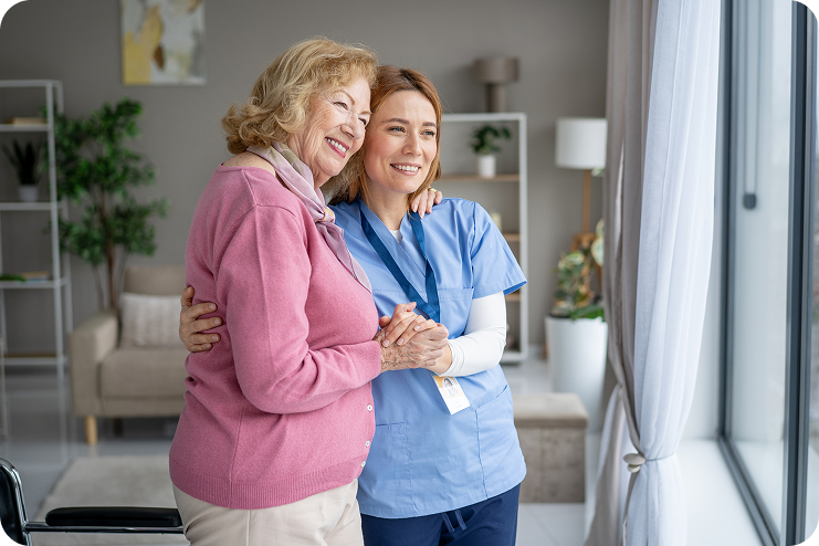 A young female caregiver in blue scrubs smiles and holds the hand of a happy elderly woman as they stand together, looking out a large window.