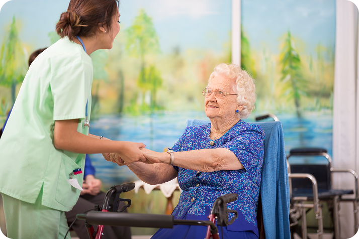 A happy elderly woman in a blue dress, sitting in a wheelchair, holds the hands of a smiling female caregiver in green scrubs.