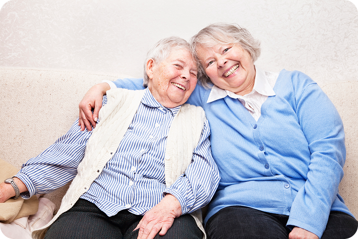 Two happy elderly women with their arms around each other, sitting close together and smiling at the camera.