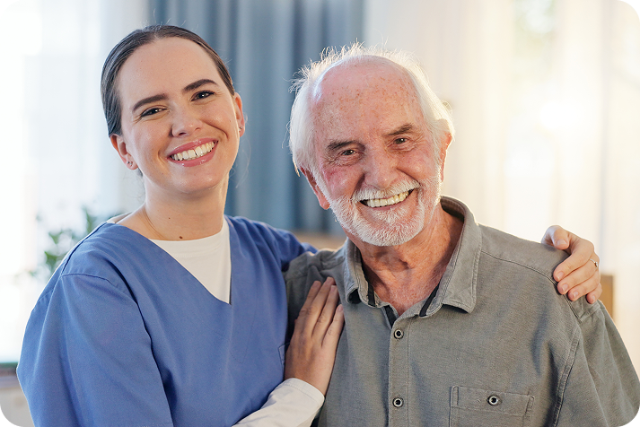 A smiling young female caregiver in blue scrubs stands with her arm around the shoulder of a happy, elderly man.