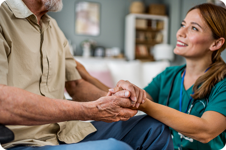 A caring female caregiver in green scrubs holds the hands of a senior man as they smile at each other.
