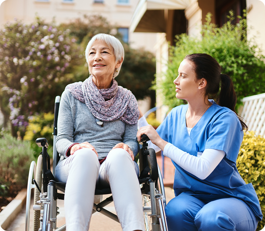 A happy senior woman in a wheelchair smiles as her caregiver kneels beside her outdoors.