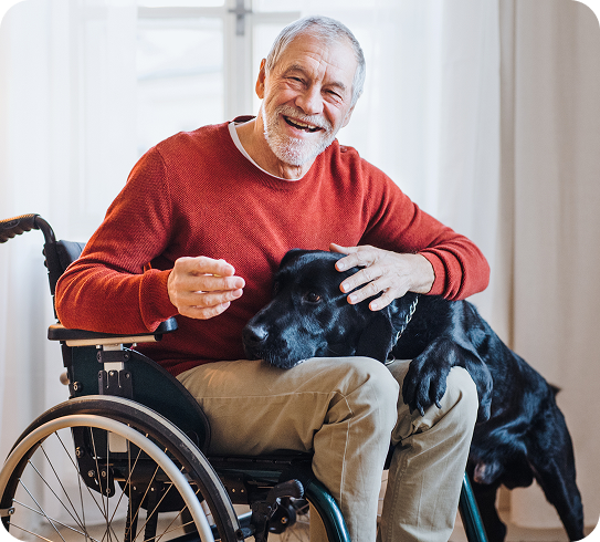 A smiling senior man in a wheelchair pets a black lab dog.