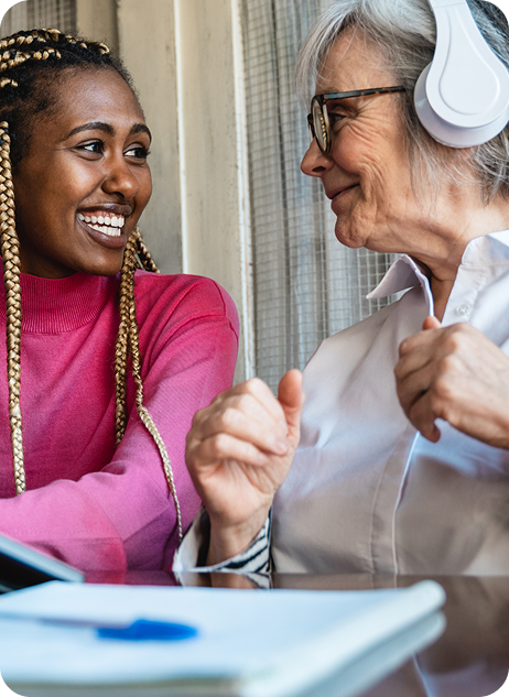 Two women, one with braids and the other with gray hair in headphones, share a joyful conversation in a bright setting.