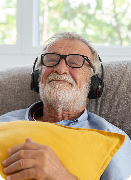 Senior man with headphones relaxes on a couch, holding a yellow pillow and smiling while listening to music.