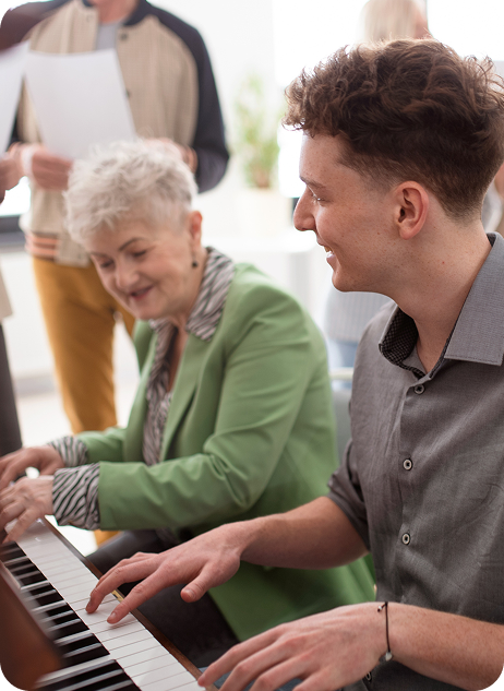Adults enjoying a music session at a piano, with one woman in a green blazer and a young man actively playing.