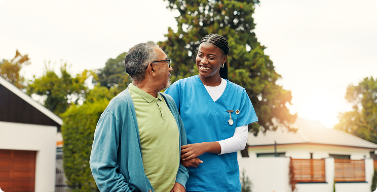 Caregiver and elderly man share a friendly conversation outdoors, with greenery and residential buildings in the background.