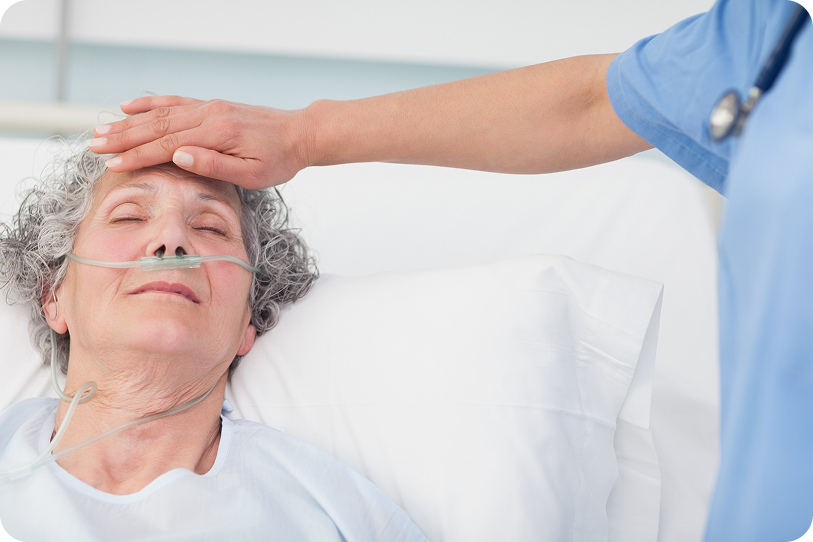 An elderly woman resting on a hospital bed receives care from a healthcare professional with a gentle touch on her forehead.