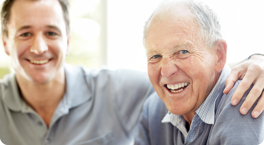 A middle-aged man smiles with an older man in a bright environment, both appearing happy and engaged in conversation.