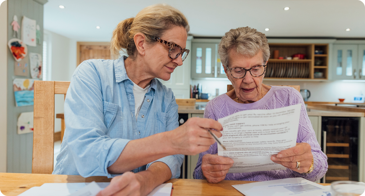 Two women review printed documents together at a wooden table in a brightly lit kitchen space.