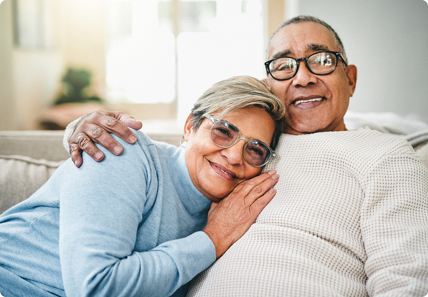 Elderly couple comfortably embracing on a sofa, smiling and enjoying a cozy moment together at home.
